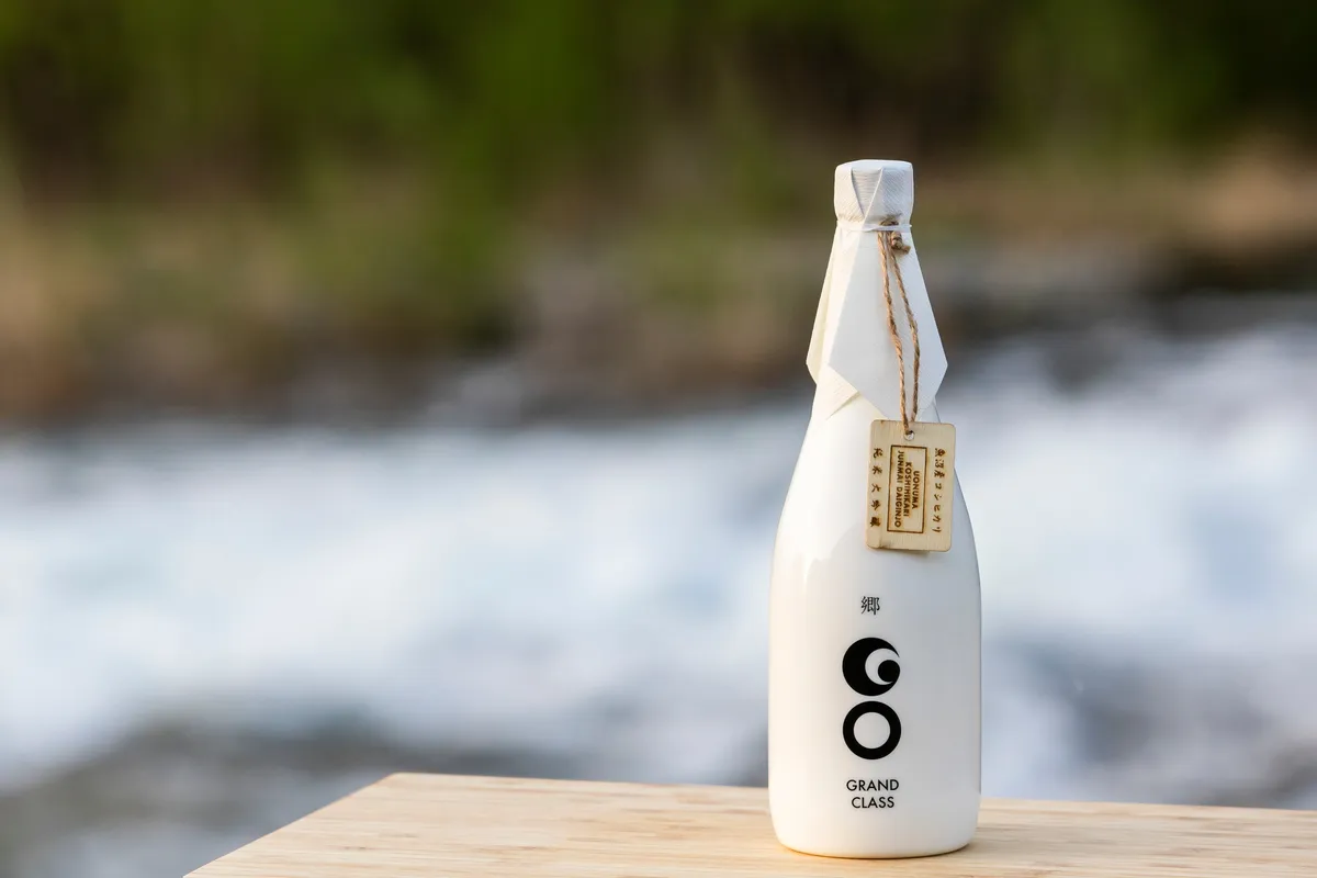 ขวด GO GRANDCLASS Uonuma Koshihikari Edition โดย Tsunan Sake Brewery บนพื้นไม้ข้างแม่น้ำที่ไหลผ่าน